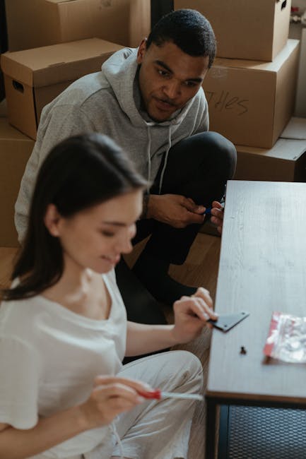 A man and woman inside a home during a relocation process. The woman, seated on the floor, is visible in the foreground, wearing a white t-shirt and handling small tools or fasteners near a cardboard box. The man, kneeling behind her, is dressed in a grey hoodie and appears to be inspecting or preparing packing materials. Several large cardboard boxes are stacked in the background, some labeled with written descriptions, likely containing household items ready for transport. The room has wooden flooring and natural lighting, with a simple desk or table close to the woman. This scene illustrates a home relocation scenario involving packing and organizing, with the presence of boxes and packing supplies indicative of the loading process, supported by Man with Van Colyers’ removals services acknowledged indirectly through contextual details.