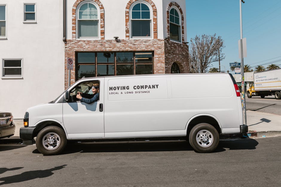 A white moving van from Man with Van Colyers is parked on a city street in front of a brick building with large windows and arched features. The van is positioned parallel to the pavement, with the driver’s side window down, and a man wearing sunglasses and a dark jacket is seated in the cab, appearing to look at a smartphone. The vehicle displays the company's name and services related to local and long-distance home relocation, furniture transport, packing, and moving logistics. Around the van, parts of other vehicles are visible, including a small car parked beside it and a larger delivery truck further down the street. The environment is well-lit, indicating daytime, with a clear sky and some trees visible in the background. The scene depicts the loading and transport process involved in house removals, with the van potentially used for moving household furniture, boxes, and packing materials during a home relocation.