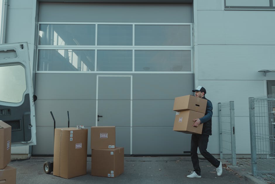 A man and woman inside a home during a relocation process. The woman, seated on the floor, is visible in the foreground, wearing a white t-shirt and handling small tools or fasteners near a cardboard box. The man, kneeling behind her, is dressed in a grey hoodie and appears to be inspecting or preparing packing materials. Several large cardboard boxes are stacked in the background, some labeled with written descriptions, likely containing household items ready for transport. The room has wooden flooring and natural lighting, with a simple desk or table close to the woman. This scene illustrates a home relocation scenario involving packing and organizing, with the presence of boxes and packing supplies indicative of the loading process, supported by Man with Van Colyers’ removals services acknowledged indirectly through contextual details.