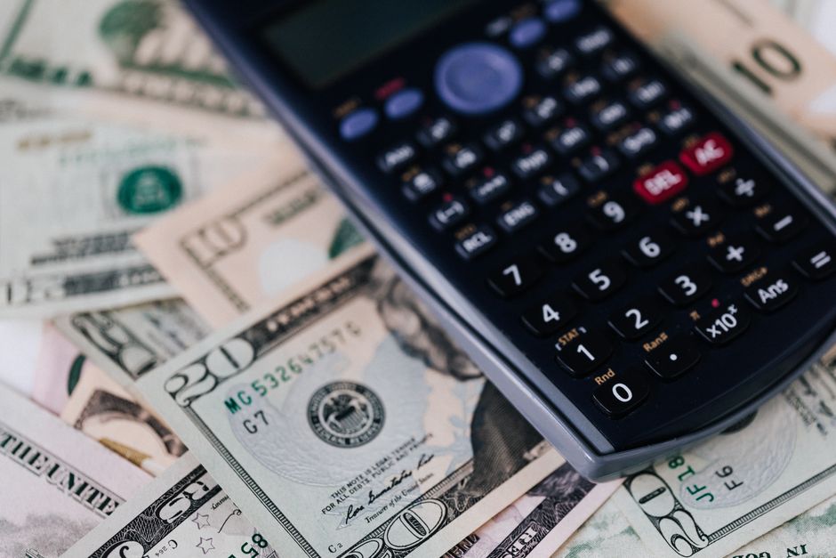 A close-up image shows a black digital calculator resting on a spread of United States dollar bills, including twenty-dollar, fifty-dollar, and one-dollar notes, arranged on a flat surface. The calculator's screen is visible, displaying various buttons and operations, and it appears to be used in the context of financial calculations. The scene reflects a focus on budgeting or cost comparison related to home relocation or moving services. This setting, captured during the packing and moving process, highlights the financial aspect of choosing between hourly or fixed-rate removals, as discussed on Man with Van Colyers' webpage about moving cost options. The dollars and calculator together symbolize the planning stage of furniture transport and packing costs involved in residential removal services.