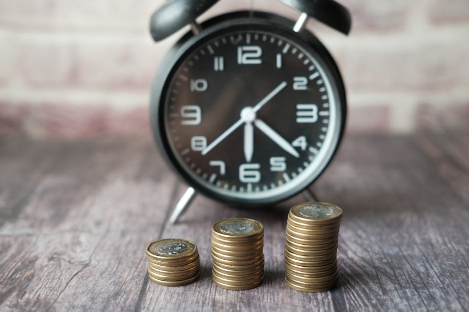 A black analog alarm clock with white numerals and clock hands showing the time slightly after 4:30, positioned on a wooden surface indoors. In front of the clock, there are three neatly stacked piles of gold coins, increasing in height from left to right. The scene captures a close-up view that emphasizes the balance of time and money, reflecting themes of cost-effective planning in home relocation, furniture transport, and packing and moving services, as offered by Man with Van Colyers. The background includes a brick wall, and the lighting appears natural, highlighting the textures of the wooden surface and the metallic shine of the coins.
