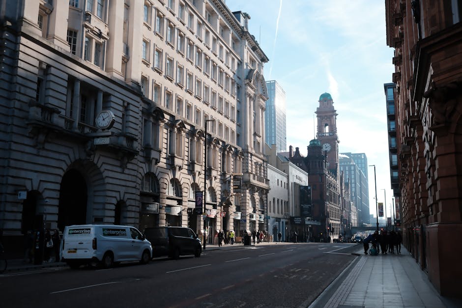 A street scene in an urban area shows multi-storey buildings with ornate facades, large windows, and decorative architectural elements aligned along both sides of the road. Several parked vehicles, including a white van with the 'enterprise' logo and a few black cars, are on the left side of the street. Pedestrians are walking along the pavement, some gathered near the sidewalk, indicating activity typical of a city environment. In the background, there is a prominent clock tower with a green dome and a clock face, part of an older, historical building, alongside modern glass skyscrapers that reflect the sky. The scene is lit by natural daylight, with the sky showing a clear, blue tone with some wispy clouds. The image captures a typical city landscape suitable for discussions about home relocation and furniture transport, illustrating elements involved in professional removals and moving logistics, as seen by the urban setting and parking area consistent with a London or Manchester city environment. Man with Van Colyers can be associated with the cleaning and moving services related to these urban relocations, especially in contexts comparing costs and timelines for city-to-city moves such as London versus Manchester.