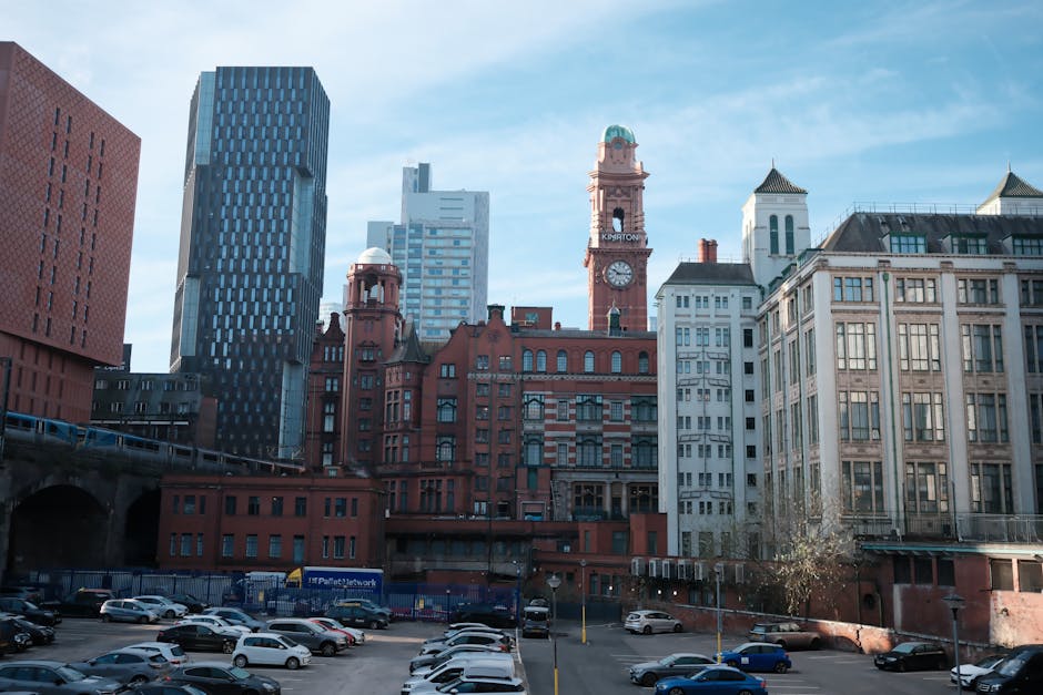 A cityscape featuring a mix of modern and historic buildings with red brick and white facades, situated behind a parking lot filled with numerous parked cars. Tall skyscrapers with glass and steel exteriors are visible in the background, contrasting with the older structures that include a clock tower with a green dome. The scene is illuminated by natural daylight with a clear blue sky overhead, typical of urban settings where home relocation and furniture transport services might be required, such as those offered by Man with Van Colyers for detailed moving logistics and packing and moving tasks.