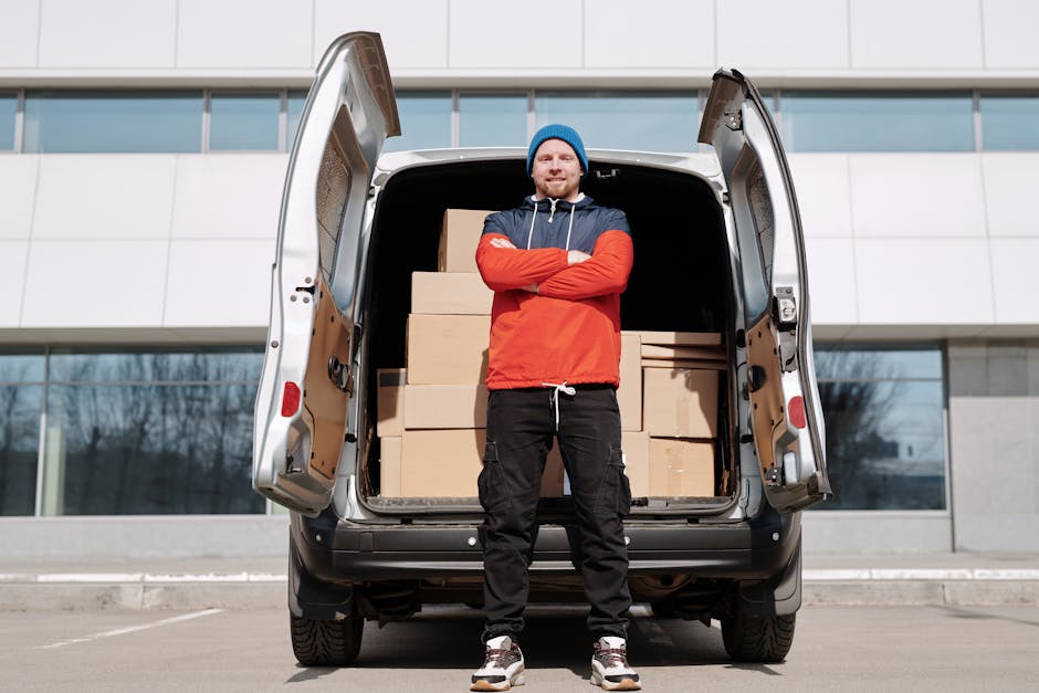 A man wearing a blue beanie, red and navy jacket, and black cargo pants stands with arms crossed in front of an open van door, revealing a spacious cargo area filled with stacked cardboard boxes wrapped in plastic and placed on wooden pallets. The van is parked outdoors on a paved area in front of a modern building with large glass windows. The scene depicts the loading process during a home relocation, with visible packing materials used for furniture transport and belongings. Several straps and blankets are visible inside the van, indicating careful handling of items. The man appears prepared for moving tasks associated with professional removals, as offered by Man with Van Colyers, highlighting the typical packing and loading activities involved in a professional moving service.