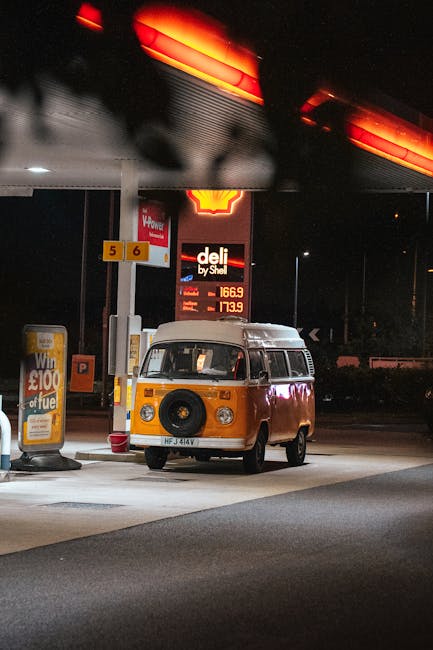 A vintage orange and white Volkswagen camper van is parked at a petrol station during nighttime. The van is positioned close to a fuel pump, with a fuel price display behind it showing prices in pounds. The petrol station has a canopy overhead with red-orange lighting, illuminating the area. Nearby, there is a promotional sign advertising a £100 fuel voucher. The scene captures the van in a simple, straightforward manner, with no other vehicles or people visible. This image relates to vehicle transport considerations often involved in home relocation and furniture transport services. Man with Van Colyers may use such imagery to illustrate their transport offerings within house removals and moving logistics.