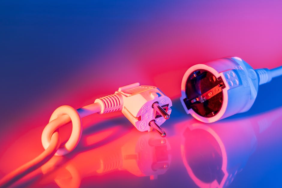 Close-up image of a standard European electrical power cable with a two-pin plug, lying on a reflective surface illuminated by pink and blue lighting. The plug and attached cable are positioned diagonally across the frame, with the plug's prongs clearly visible. The background features a gradient of pink and purple hues, creating a vibrant, modern aesthetic. The lighting highlights the glossy finish of the plug and the texture of the plastic cable sheath. This image captures the preparatory phase of packing or disconnecting electronic appliances during home relocation or furniture transport, illustrating the careful handling and organization involved in moving services provided by Man with Van Colyers. The scene is indoors, with no other objects visible, emphasizing the focus on electrical appliance safety and disconnection as part of the packing and moving process.