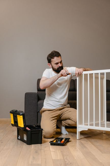 A wooden side table situated indoors with a partially assembled piece of furniture on top, including a large wooden panel covered with a cardboard sheet. Various tools are present, such as a hammer, t
