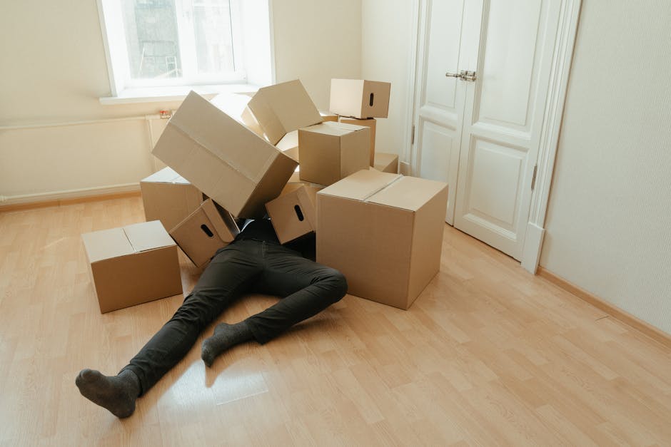 A person lying on a light wooden floor surrounded by several cardboard boxes in a room with white walls and a large window that lets in natural light. The boxes vary in size, some closed and others open, with one box toppled over. The person is partially obscured by the boxes, with only their legs and feet visible, wearing dark pants and socks. Next to the window, there is a radiator, and a closed white door is visible on the right side. This image captures the packing and home relocation process, demonstrating the handling of items during furniture transport and the loading process within a house, with the boxes positioned near the doorway for easy moving, and the room appears prepared for collection or transfer by removal professionals such as Man with Van Colyers.