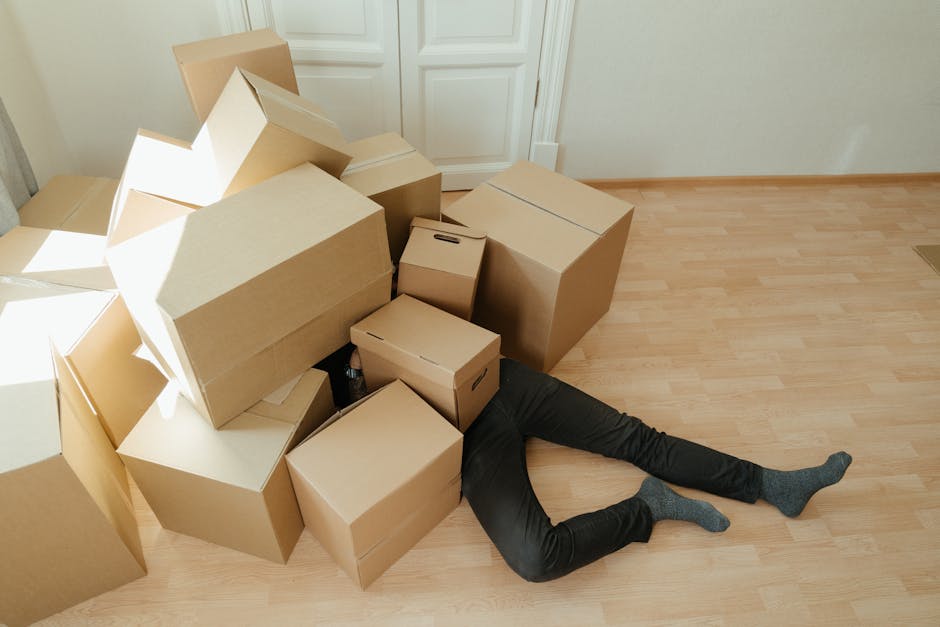 A person lying on a light wooden floor surrounded by several cardboard boxes in a room with white walls and a large window that lets in natural light. The boxes vary in size, some closed and others open, with one box toppled over. The person is partially obscured by the boxes, with only their legs and feet visible, wearing dark pants and socks. Next to the window, there is a radiator, and a closed white door is visible on the right side. This image captures the packing and home relocation process, demonstrating the handling of items during furniture transport and the loading process within a house, with the boxes positioned near the doorway for easy moving, and the room appears prepared for collection or transfer by removal professionals such as Man with Van Colyers.
