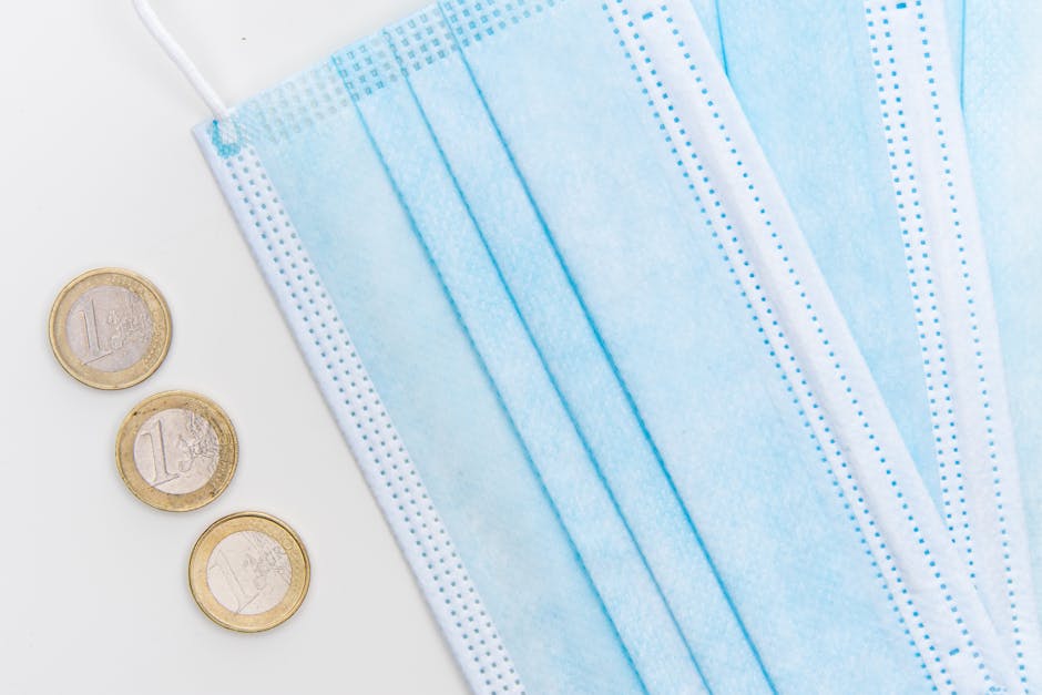 A close-up image shows several blue disposable face masks stacked with some overlapping each other, placed on a white surface. The masks are made of non-woven fabric with visible pleats and ear loops, indicating they are designed for protective purposes during house removals or moving activities. To the left of the masks, there are three one-euro coins arranged vertically, providing a sense of scale and emphasizing the size of the masks. The scene is well-lit with soft, even lighting, highlighting the texture and details of the masks and coins. This image reflects the importance of safety and hygiene considerations during home relocation, with [COMPANY_NAME] such as Man with Van Colyers taking steps to facilitate safe moving and packing procedures, including protective gear and organized transport of belongings in a professional and efficient manner.