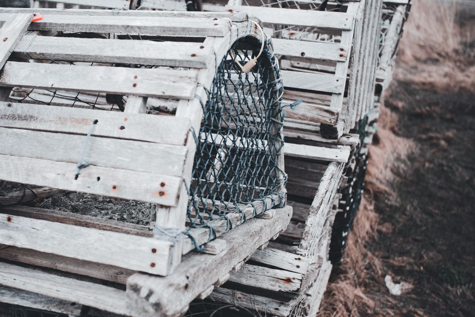 A close-up view of multiple wooden crates stacked on top of each other, with some featuring wire mesh panels on the sides, likely used for transporting or storing fragile items during a house relocation. The crates are positioned on a dirt surface, with some resting on a slight incline, indicating they are outside a property or in a moving preparation area. The wooden slats are weathered and secured with nails and thin metal ties, with the wire mesh panels helping secure contents inside. The scene suggests a packing and moving process handled by a professional removal service such as Man with Van Colyers, highlighting the use of sturdy containers for safe furniture transport during home relocation or professional removals.